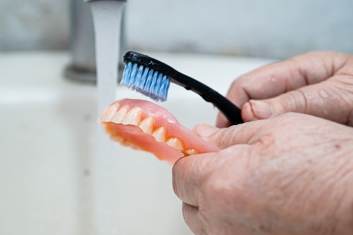 Older person holding dentures over a sink and cleaning them with a brush