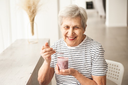An elderly woman smiles while eating yogurt.