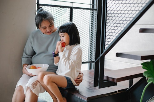 Asian woman giving fruits to hungry granddaughter at home