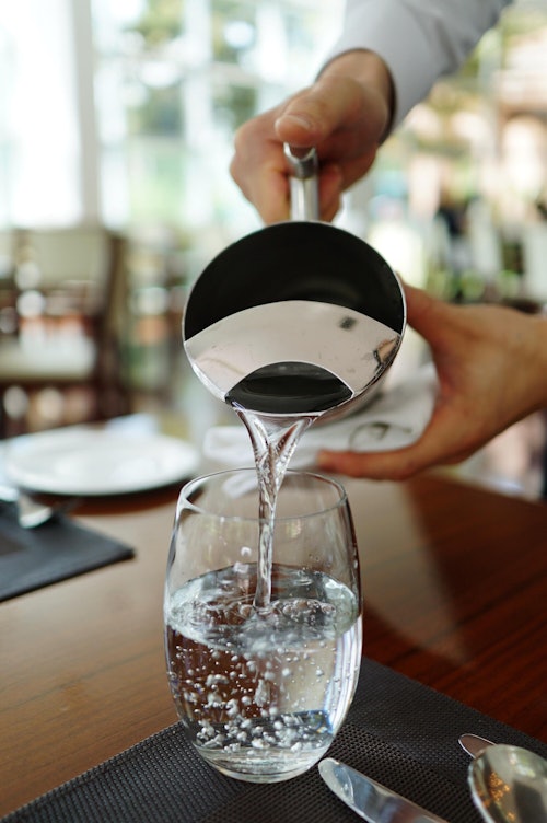 person pouring water into glass from metal pitcher